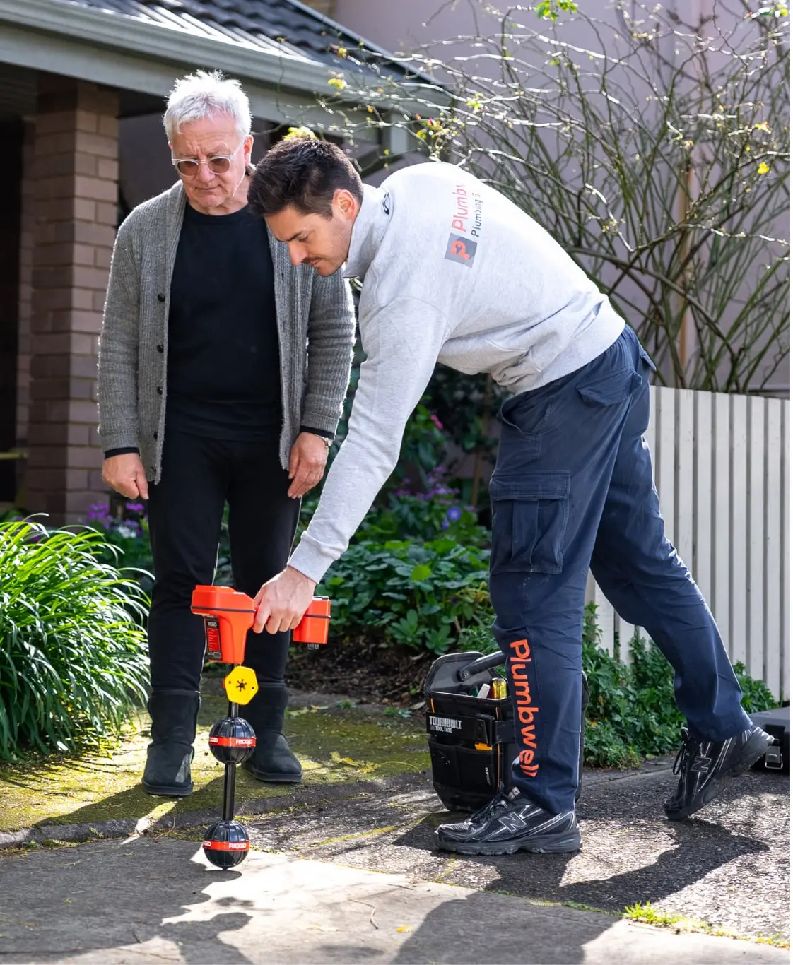 Plumber using a pipe locator on a front path while the owner watches